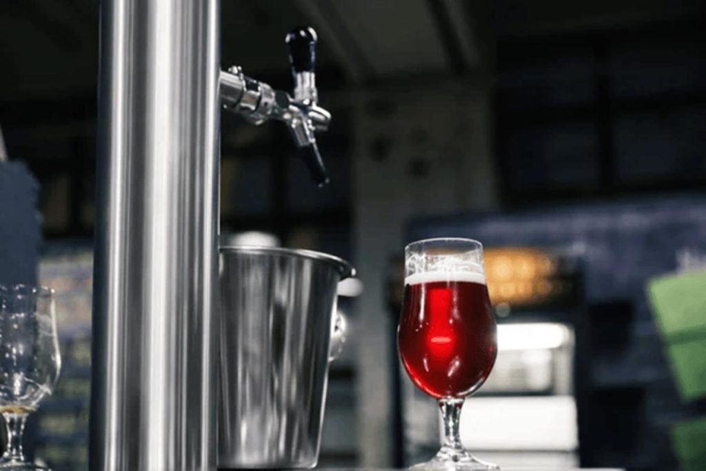 A freshly poured craft beer, served in a glass beside a stainless steel draft tap in a brewery setting