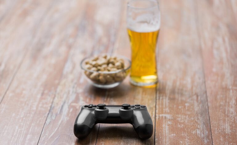 Glass of craft beer, game controller, and bowl of snacks on rustic wooden table
