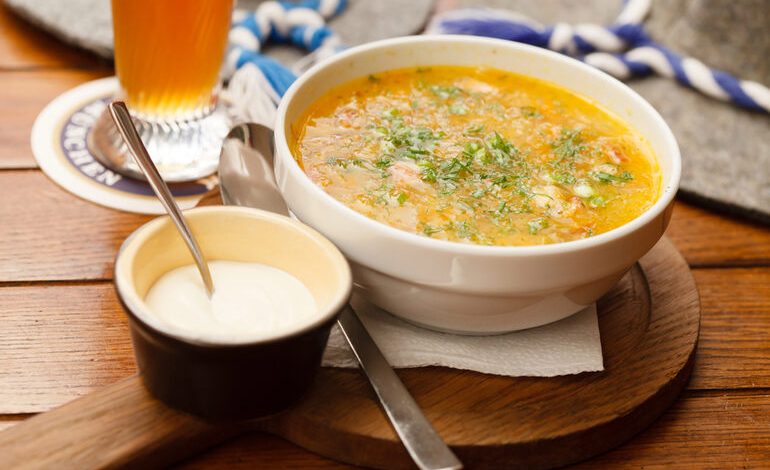 A hearty bowl of beer cheese soup garnished with herbs, served with a side of sour cream and a glass of beer