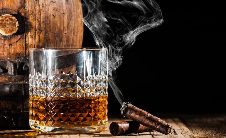 A glass of whiskey with smoke rising from cigars beside a wooden barrel