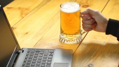 Person holding a glass mug of beer while using a laptop on a wooden table, the intersection of brewing and digital technology