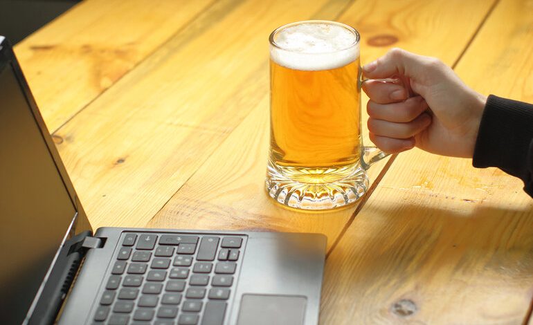 Person holding a glass mug of beer while using a laptop on a wooden table, the intersection of brewing and digital technology