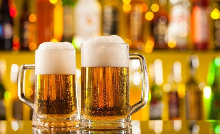 Two frothy pints of craft beer on a bar counter with colorful bottles in the background, perfect for enjoying while playing slots