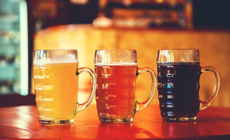 Three mugs of craft beer in varying colors, from light to dark, on a bar counter in a cozy, dimly lit San Diego taproom, perfect for a late-night drink