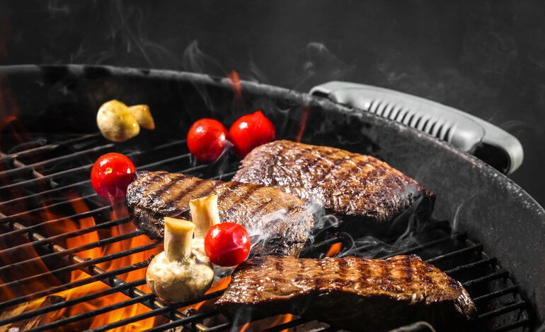 Grilled steaks, cherry tomatoes, and mushrooms cooking over open flames on a charcoal BBQ grill with visible smoke