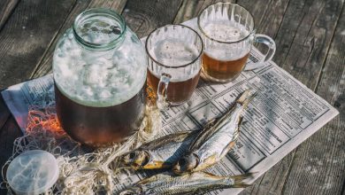 Rustic setup with frothy beer in a glass jar and mugs served alongside dried fish on newspaper