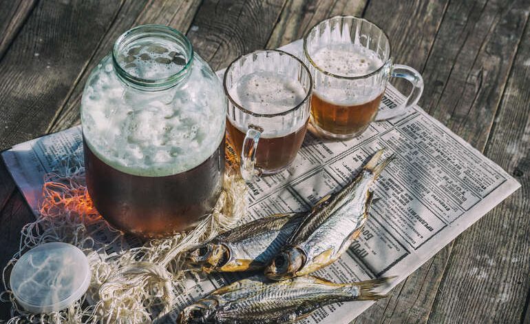Rustic setup with frothy beer in a glass jar and mugs served alongside dried fish on newspaper