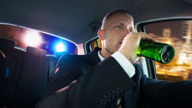 Man drinking beer while driving as police car lights are visible in the background during a DUI traffic stop.