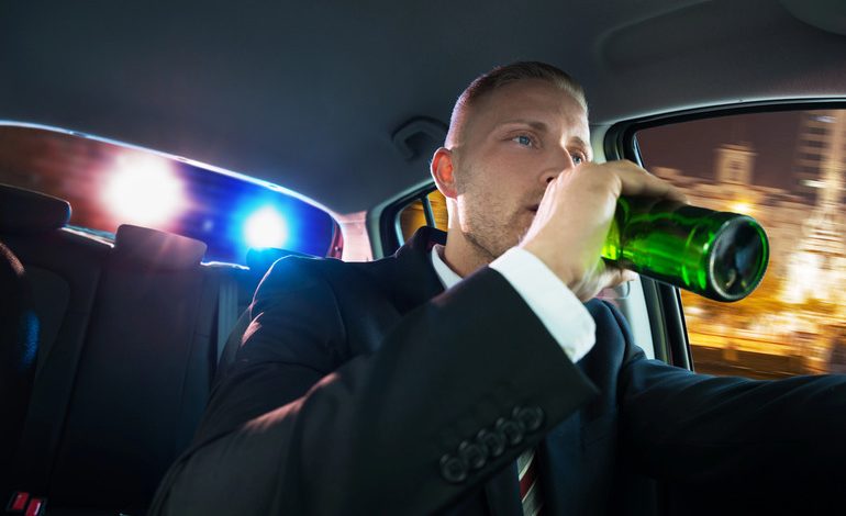 Man drinking beer while driving as police car lights are visible in the background during a DUI traffic stop.