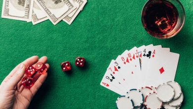 A casino gaming table with red dice, playing cards, poker chips, stacks of hundred-dollar bills, and a glass of whiskey on a green felt surface.