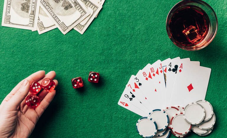A casino gaming table with red dice, playing cards, poker chips, stacks of hundred-dollar bills, and a glass of whiskey on a green felt surface.