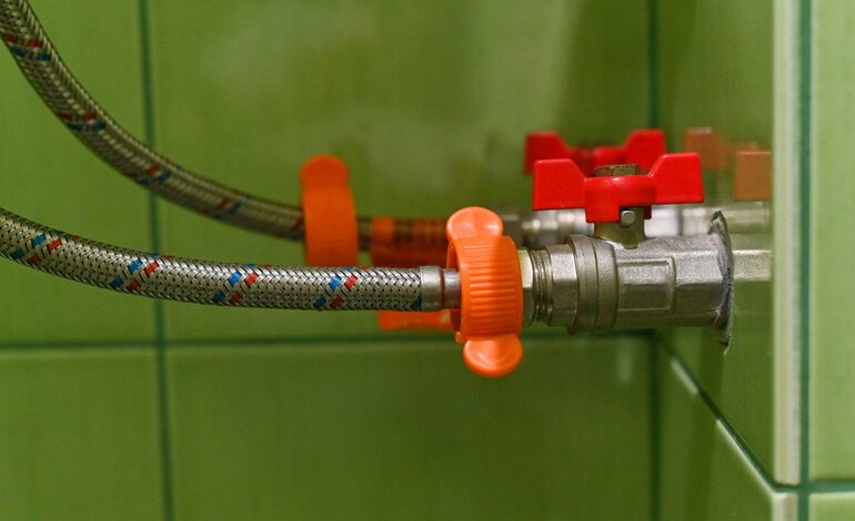 Close-up of a braided steel drain hose connected to a wall valve with red and orange knobs, demonstrating proactive drain care setup for brewers.