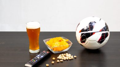 Glass of craft beer, potato chips, remote control, pistachios, and a soccer ball on a table, ideal game day snacks and drinks setup.