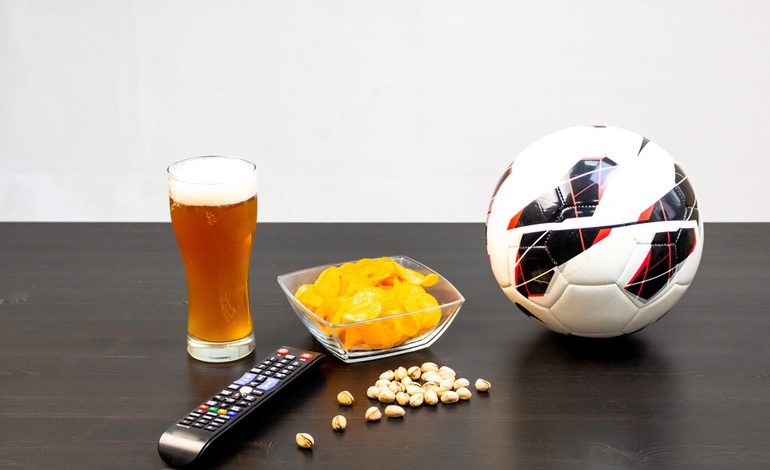 Glass of craft beer, potato chips, remote control, pistachios, and a soccer ball on a table, ideal game day snacks and drinks setup.