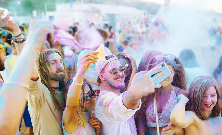 A Group of friends covered in colorful powder, taking a selfie, and celebrating at an outdoor festival, enjoying vibrant energy and summer vibes.