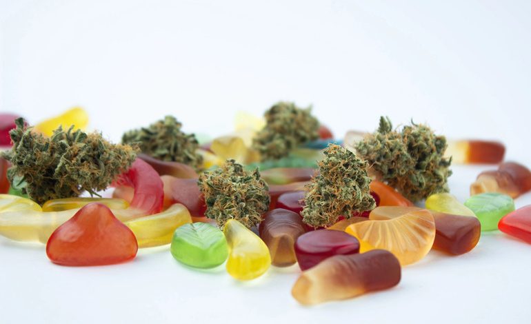 Assorted colorful gummy candies scattered with several cannabis buds on a white background.