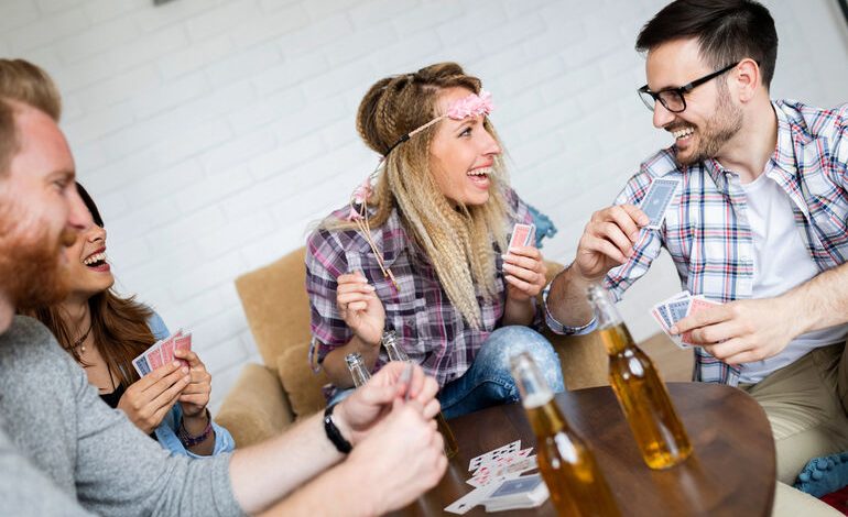 Friends laughing and playing a card game together at a bar table with beer bottles.