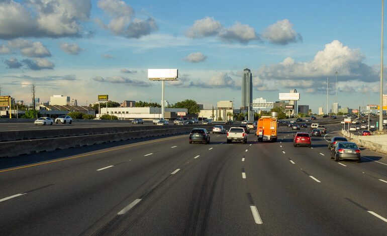 Wide highway with multiple lanes, cars, and an HOV lane during daytime under a partly cloudy sky.