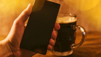 Hand holding a smartphone next to a glass of dark beer on a wooden table, symbolizing digital payments and mobile e-wallet use.