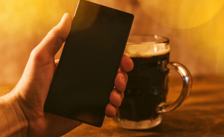 Hand holding a smartphone next to a glass of dark beer on a wooden table, symbolizing digital payments and mobile e-wallet use.