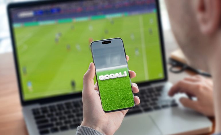Person using a mobile sports platform while watching a live football match on a laptop, illustrating digital fan engagement in Southeast Asia.
