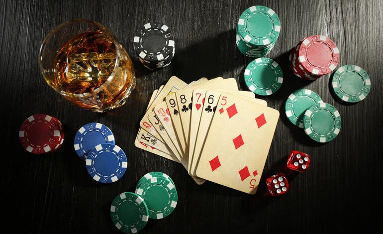 Poker cards, casino chips, dice, and a glass of alcohol on a dark table, which depicts the connection between gambling and drinking alcohol.