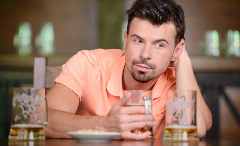 Man sitting in a bar holding a beer glass, other beer mugs on the table, looking pensive.