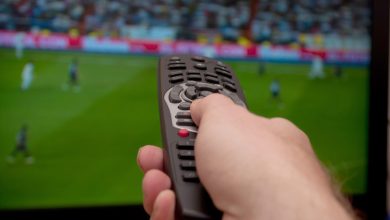 A man is holding a TV remote while watching a soccer game on the TV.