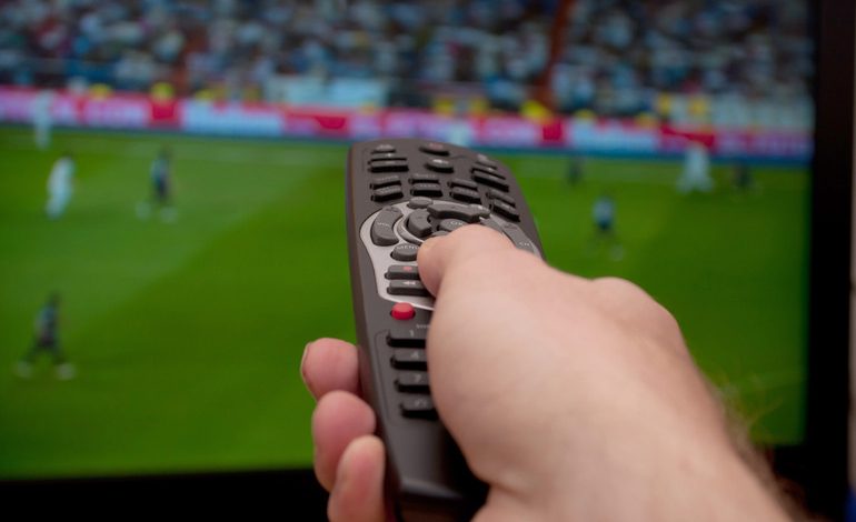 A man is holding a TV remote while watching a soccer game on the TV.