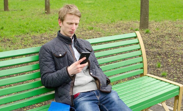 A man on a park bench checking his phone, representing how social media algorithms feed content to beer fans.
