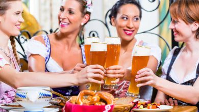 Four women in traditional dirndls clinking tall glasses of beer over a table with pretzels and food.