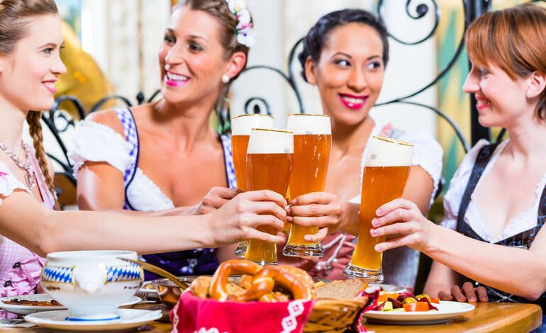 Four women in traditional dirndls clinking tall glasses of beer over a table with pretzels and food.