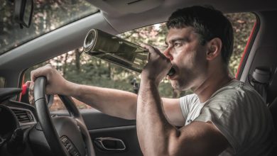 Man driving a car while drinking from a glass bottle, holding the steering wheel with one hand.