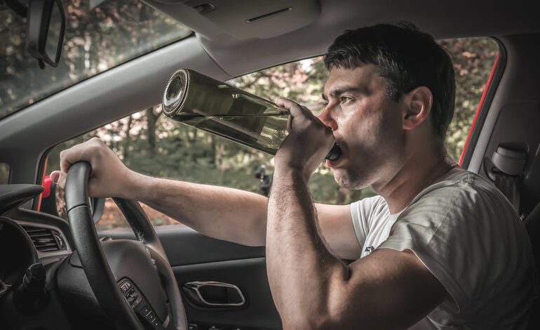 Man driving a car while drinking from a glass bottle, holding the steering wheel with one hand.