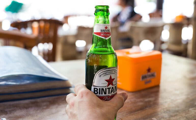 Hand holding a cold bottle of Bintang beer on a wooden table in an Indonesian café.