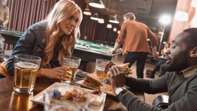 A couple is enjoying beers and sandwiches at a bar while playing pool in the background.