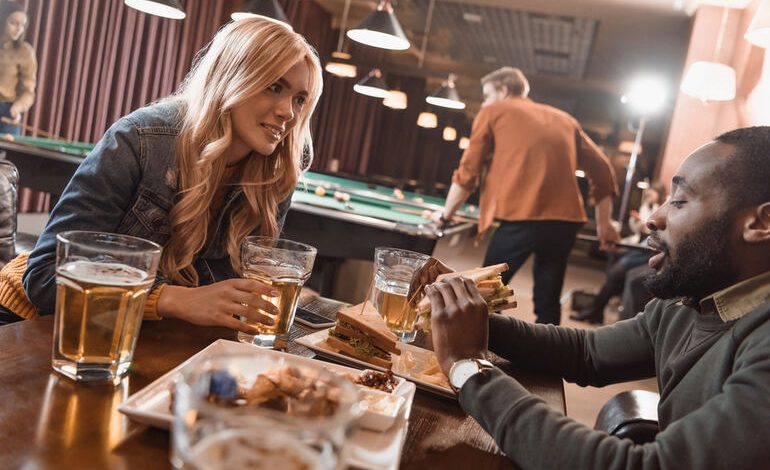 A couple is enjoying beers and sandwiches at a bar while playing pool in the background.