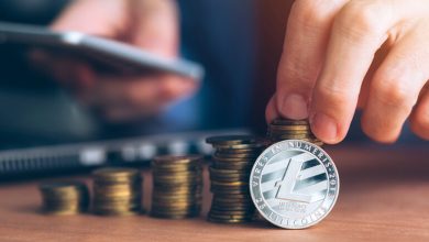 Hand stacking coins with a Litecoin coin in front and a smartphone in the background.