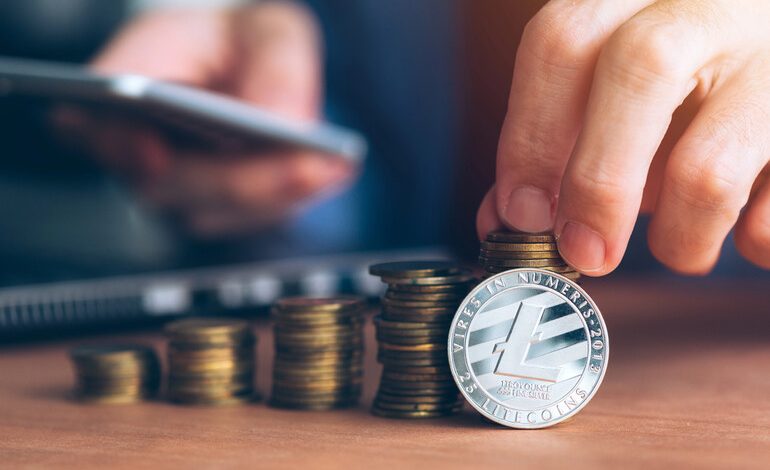Hand stacking coins with a Litecoin coin in front and a smartphone in the background.