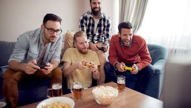 Four friends sitting on a couch playing a competitive video game with controllers, beers, and snacks on the table.