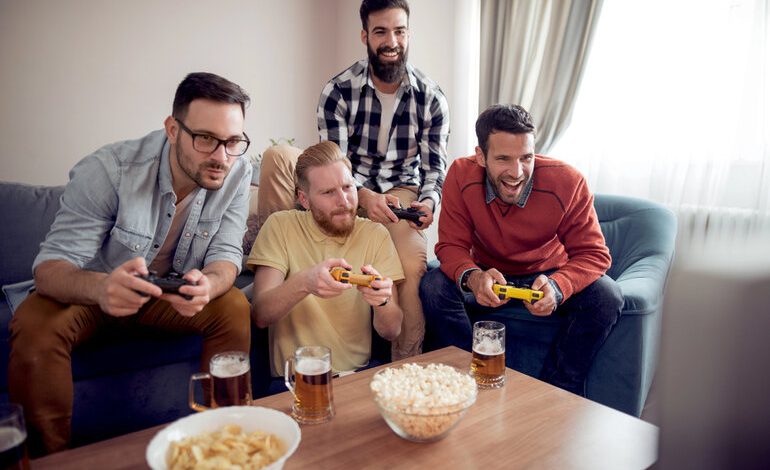 Four friends sitting on a couch playing a competitive video game with controllers, beers, and snacks on the table.