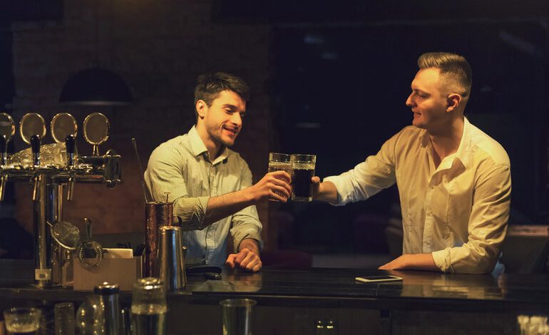 Two friends clinking glasses of beer while chatting at a cozy bar counter in the evening.