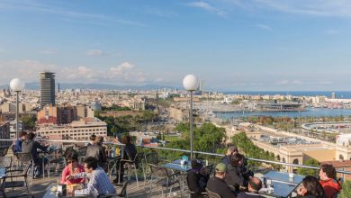 People enjoying drinks at a sunny rooftop bar overlooking a coastal city harbor and mountains.