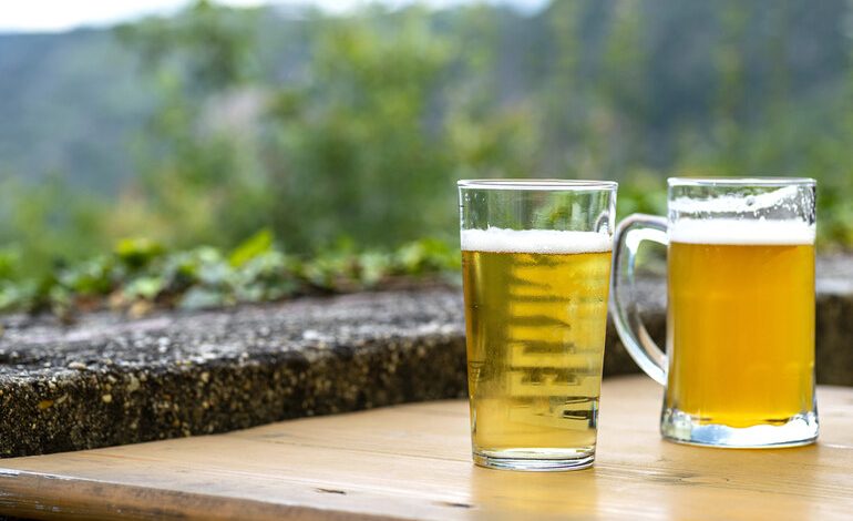 Two cold beers in clear glass mugs resting on a wooden table with a scenic green mountain backdrop.