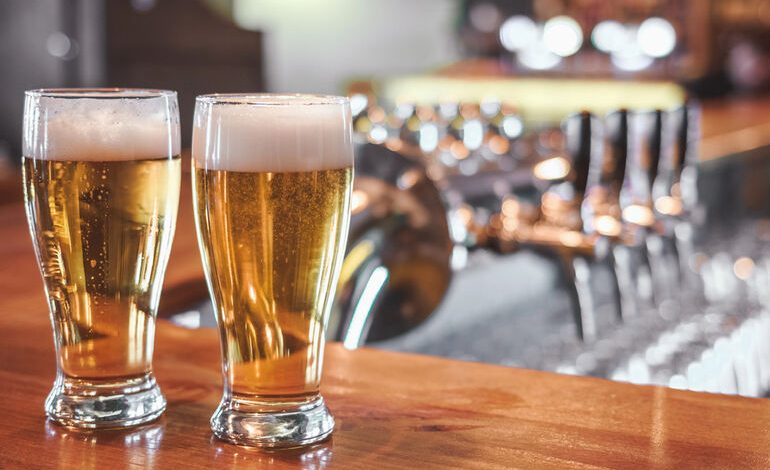 Two golden craft beers on a bar counter with taps in the background.